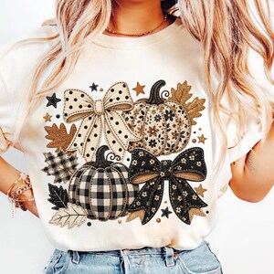 May include: A cream-colored t-shirt featuring a graphic print of two pumpkins and two bows. One pumpkin has a floral pattern, the other a black and white buffalo check. The bows are polka-dotted and floral. Brown and black leaves and stars surround them.