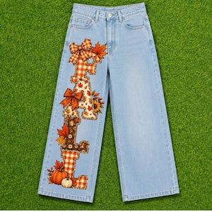 May include: Light blue denim jeans featuring a vertical graphic on the left leg that spells out the word FALL. The letters are decorated with orange and white plaid patterns, autumn leaves, orange bows, and small pumpkins at the base.