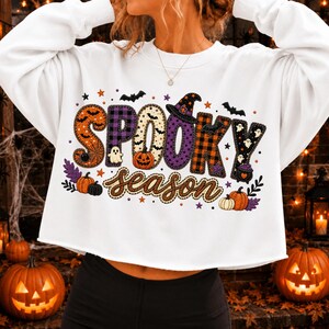 May include: A white long-sleeve cropped sweatshirt featuring the text SPOOKY season in colorful patterned letters. The design includes orange pumpkins, purple and orange plaid, black bats, small ghosts, a witch hat, and a cauldron graphic.