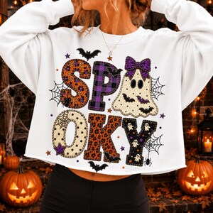 May include: A white long-sleeve sweatshirt featuring the word SPOOKY in large, patterned letters. The S is orange floral, the P is purple plaid, the O is cream with spiderwebs, the O is orange leopard print, the K is black with ghosts, and the Y is black with ghosts. A ghost with a purple bow is included.