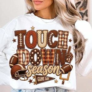 May include: A white crewneck sweatshirt featuring a graphic print with the text TOUCH DOWN season and GAME DAY. The design includes brown footballs, a football helmet, a goalpost, and a foam finger, all styled with plaid and polka dot patterns.