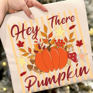 Op de afbeelding: Een beige t-shirt met een grafische print met de tekst Hey There Pumpkin in donkerrood schrift. Het ontwerp bevat een grote oranje pompoen, herfstbladeren, rode bessen, bruine paddenstoelen en gele sterren op een lichtoranje gestreepte achtergrond.