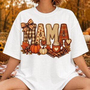 May include: A white t-shirt featuring the word MAMA in large, textured letters with plaid, knit, and leaf patterns. The design includes a plaid bow, two pumpkins, autumn leaves, and a mug of hot beverage with a heart shape on the front.