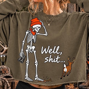 May include: An olive green long-sleeve sweatshirt featuring a graphic of a skeleton wearing an orange beanie and red scarf, holding binoculars. The skeleton stands near a deer, with the text "Well, shit..." printed in white script.