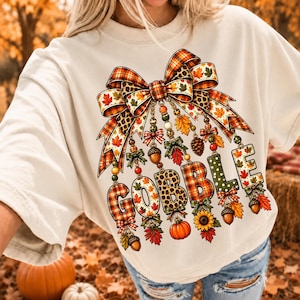 May include: A cream-colored t-shirt featuring a large graphic of a decorative bow with hanging elements. The design includes the word GOBBLE spelled out in letters decorated with plaid, leopard print, and autumn leaves, accented by pumpkins and acorns.