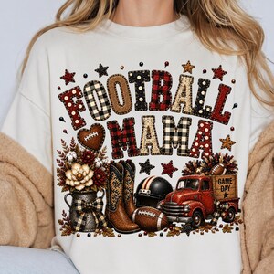 May include: A cream-colored sweatshirt featuring a graphic print with the text FOOTBALL MAMA in patterned letters. The design includes a floral arrangement in a pitcher, brown cowboy boots, a football helmet, a football, and a red truck.