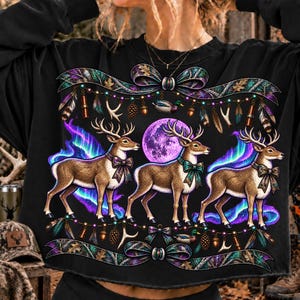 Puede incluir: Una sudadera negra de manga larga con un estampado gráfico de tres ciervos marrones con astas, cada uno con un lazo. El diseño incluye una luna morada, auroras boreales azules y moradas, plumas colgantes, piñas y cintas decorativas.