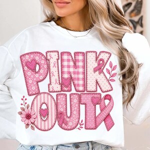 May include: A white long-sleeve sweatshirt featuring the text PINK OUT in large, block-style letters. Each letter is filled with various pink patterns, including gingham, polka dots, stripes, and floral motifs, accented with small hearts and ribbons.