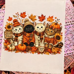 May include: A white fabric surface featuring a graphic illustration of four kittens wearing autumn-themed clothing, scarves, and hats. The kittens are surrounded by orange pumpkins, sunflowers, and fall leaves. A sign reads HELLO FALL and a mug reads PUMPKIN SPICE.