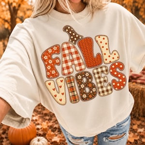 May include: An off-white crewneck t-shirt featuring a large graphic on the chest that reads FALL VIBES. The letters are filled with various patterns including red and white gingham, orange knit textures, floral prints, and polka dots.