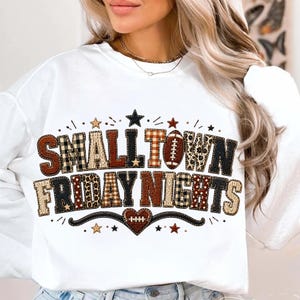 May include: A white long-sleeve sweatshirt featuring the text "SMALL TOWN FRIDAY NIGHTS" in a multi-patterned font. The letters incorporate brown, black, and white plaid, leopard print, and knit textures, with a football and heart graphic.