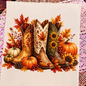 Può includere: Una stampa su tela rettangolare che raffigura tre stivali da cowboy disposti insieme. Gli stivali sono decorati con motivi autunnali tra cui foglie d'acero, girasoli e bacche. Diverse zucche arancioni e bianche e pigne circondano gli stivali.