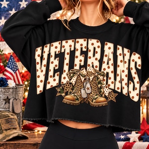 Puede incluir: Una sudadera negra de manga larga y corte corto con el texto VETERANS en letras grandes de color crema con lunares marrones. Debajo del texto hay un gráfico de dos botas de combate de camuflaje con lazos, placas de identificación y estrellas.