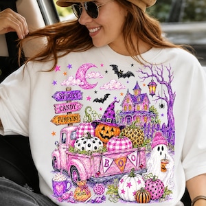 May include: A white sweatshirt featuring a colorful Halloween graphic. The design includes a pink truck carrying pumpkins, a haunted house, a ghost, bats, a crescent moon, and signs reading SPOOKY, CANDY, and PUMPKINS, with a BOO banner on the truck.