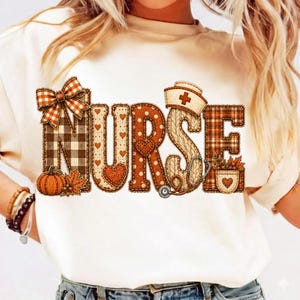 May include: A cream-colored t-shirt featuring the word NURSE in large, decorative letters. The letters are filled with autumn-themed patterns including plaid, polka dots, and hearts, accented by a pumpkin, a bow, and a stethoscope graphic.