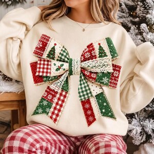 May include: A cream-colored long-sleeve sweatshirt featuring a large, central graphic of a decorative bow. The bow is composed of various fabric patterns including red and green plaid, white snowflakes on red, green trees, and lace trim.