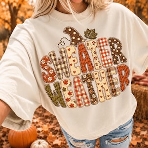 May include: A cream-colored short-sleeve t-shirt featuring the text "SWEATER WEATHER" in large, blocky letters. Each letter is filled with a different autumn-themed pattern, including plaid, polka dots, hearts, leaves, and floral prints in warm tones.