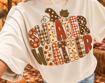 May include: A cream-colored short-sleeve t-shirt featuring the text "SWEATER WEATHER" in large, blocky letters. Each letter is filled with a different autumn-themed pattern, including plaid, polka dots, hearts, leaves, and floral prints in warm tones.