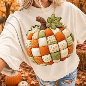 May include: A cream-colored short-sleeve t-shirt featuring a large, three-dimensional quilted pumpkin graphic. The pumpkin is constructed from a patchwork of orange, cream, and green fabrics with floral and gingham patterns, topped with a brown stem.
