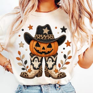 May include: A cream-coloured t-shirt featuring a graphic of an orange jack-o'-lantern wearing a black cowboy hat with leopard print trim. Below the pumpkin are two beige cowboy boots decorated with white ghost figures and leopard print accents.