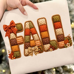 Puede incluir: Una superficie de tela blanca con la palabra FALL escrita en letras acolchadas tridimensionales de estilo patchwork. Las letras están compuestas por varios cuadrados de tela en colores otoñales naranja, marrón, verde y crema, decoradas con lazos y hojas.