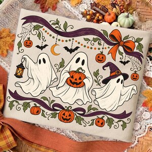 May include: A rectangular fabric panel featuring a cream background with a Halloween illustration. Three white ghosts are depicted: one holding a lantern, one holding a jack-o'-lantern, and one wearing a purple witch hat. Orange pumpkins, bats, and purple ribbons decorate the scene.