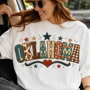 May include: A white crewneck sweatshirt featuring the word OKLAHOMA in large block letters. Each letter has a unique pattern including floral, plaid, and geometric designs. The graphic is accented with small stars and a central red heart.
