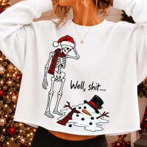 Könnte beinhalten: Ein weißes Langarm-Sweatshirt mit einer Grafik eines Skeletts, das eine rote Weihnachtsmütze und einen rot karierten Schal trägt und über einem geschmolzenen Schneemann mit schwarzem Zylinder steht, inklusive dem schwarzen Schriftzug "Well, shit..."