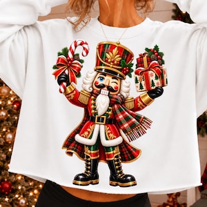 Puede incluir: Una sudadera blanca de manga larga con el gráfico de un cascanueces con uniforme de cuadros rojos y verdes, sosteniendo un bastón de caramelo y un regalo envuelto, con barba blanca y botas negras.
