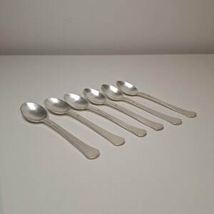 Könnte beinhalten: Ein Set aus sechs silberfarbenen Metalllöffeln, die in einer diagonalen Reihe angeordnet sind. Jeder Löffel hat eine glatte, abgerundete Laffe und einen schlichten, flachen Stiel, der sich zum Ende hin leicht verjüngt. Polierte, glänzende Oberfläche.