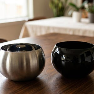 May include: Two spherical bowls sit side by side. One bowl features a brushed stainless steel finish with a metallic silver tone, while the other bowl has a smooth, glossy black finish. Both containers have a rounded, deep shape with open tops.