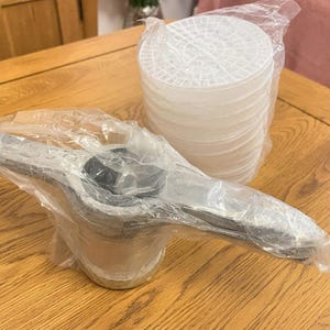 May include: A silver-colored metal dishwasher spray arm with a central hub and two extending arms, encased in clear plastic packaging, accompanied by a stack of white circular plastic dishwasher filter inserts also wrapped in clear plastic.