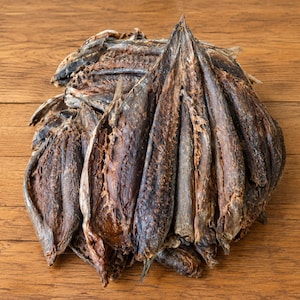 May include: A pile of dried, dark brown fish fillets arranged in a fan-like formation. Each piece of fish has a textured, leathery appearance with visible skin and flesh, showing a deep, concentrated color throughout the dried seafood product.