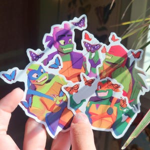 Puede incluir: Una pegatina troquelada que muestra cuatro personajes de tortugas de dibujos animados con piel verde y petos naranjas, cada uno con una máscara de diferente color. Los personajes están rodeados de mariposas coloridas en tonos morados, azules y naranjas.