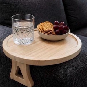 May include: A round, light-colored wooden sofa arm tray with a flat surface and two side supports that grip the armrest. On the tray sits a clear, textured glass filled with water and a small, light-colored bowl containing crackers and grapes.