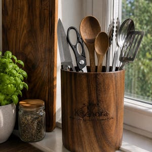 May include: A cylindrical dark wood utensil holder with the engraved text Kitchora COOK CREATE GATHER. It contains a metal knife, black kitchen shears, two wooden spoons, metal tongs, and a metal slotted spatula, all standing upright.