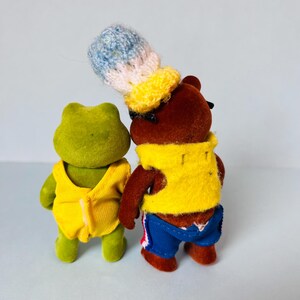 May include: A green flocked frog figurine wearing a yellow sleeveless top and a brown flocked bear figurine wearing a yellow vest, blue shorts with red accents, and a tall, textured, multi-colored knit hat in blue, white, and yellow.