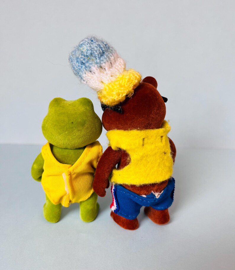 May include: A green flocked frog figurine wearing a yellow sleeveless top and a brown flocked bear figurine wearing a yellow vest, blue shorts with red accents, and a tall, textured, multi-colored knit hat in blue, white, and yellow.