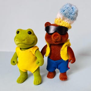 May include: A green frog figurine wearing a yellow sleeveless top stands next to a brown bear figurine. The bear wears black sunglasses, a yellow vest, blue shorts with red side stripes, and a blue and white knitted hat on its head.