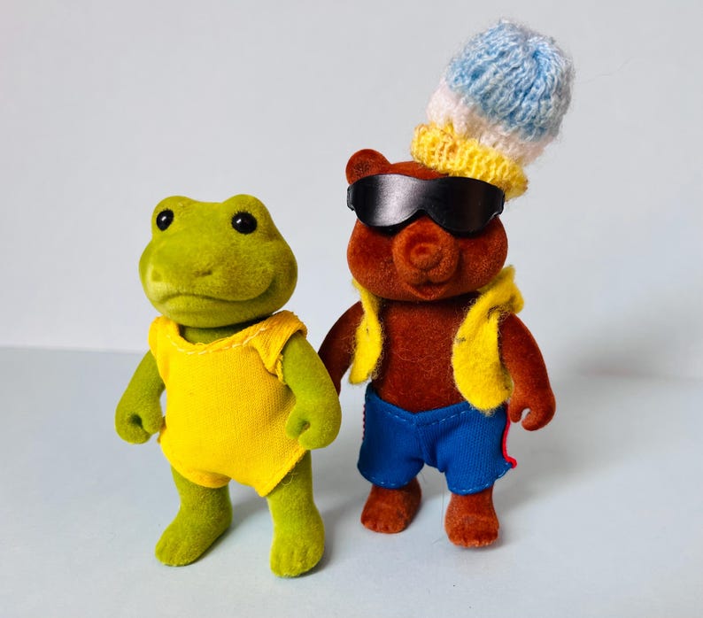 May include: A green frog figurine wearing a yellow sleeveless top stands next to a brown bear figurine. The bear wears black sunglasses, a yellow vest, blue shorts with red side stripes, and a blue and white knitted hat on its head.