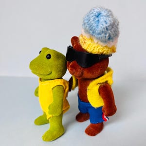 May include: A green frog figurine wearing a yellow sleeveless top stands next to a brown bear figurine. The bear wears black sunglasses, a yellow vest, blue shorts, and a multi-colored knit hat with blue, white, and yellow horizontal stripes.