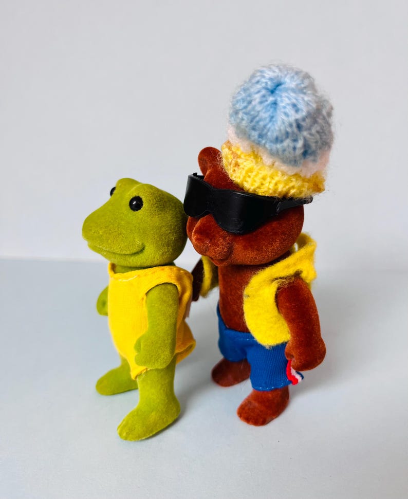 May include: A green frog figurine wearing a yellow sleeveless top stands next to a brown bear figurine. The bear wears black sunglasses, a yellow vest, blue shorts, and a multi-colored knit hat with blue, white, and yellow horizontal stripes.