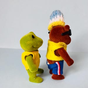 May include: A green frog figurine wearing a yellow sleeveless top stands next to a brown bear figurine. The bear wears a yellow sleeveless top, blue shorts with red and white trim, black sunglasses, and a blue, white, and yellow knit hat.