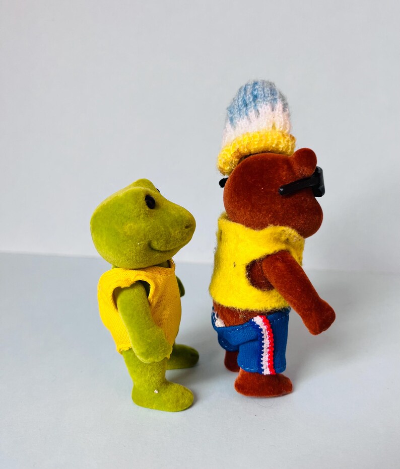 May include: A green frog figurine wearing a yellow sleeveless top stands next to a brown bear figurine. The bear wears a yellow sleeveless top, blue shorts with red and white trim, black sunglasses, and a blue, white, and yellow knit hat.