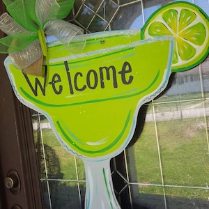 May include: A green and white painted wooden margarita glass with a lime wedge and the word "Welcome" on the front. The glass is hanging on a door with a green and silver bow.