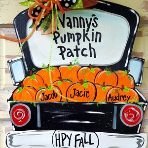 May include: Wooden sign shaped like a black truck bed filled with orange pumpkins. The sign reads "Nanny's Pumpkin Patch" and "(HPY FALL)". The pumpkins are labeled with names. A decorative bow tops the sign.