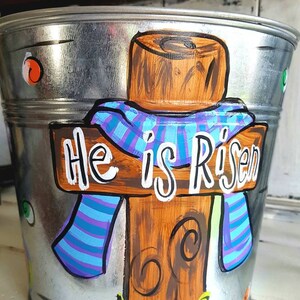 May include: A silver metal bucket painted with a wooden cross, a blue and purple scarf, and the words "He is Risen". The cross is surrounded by green grass.