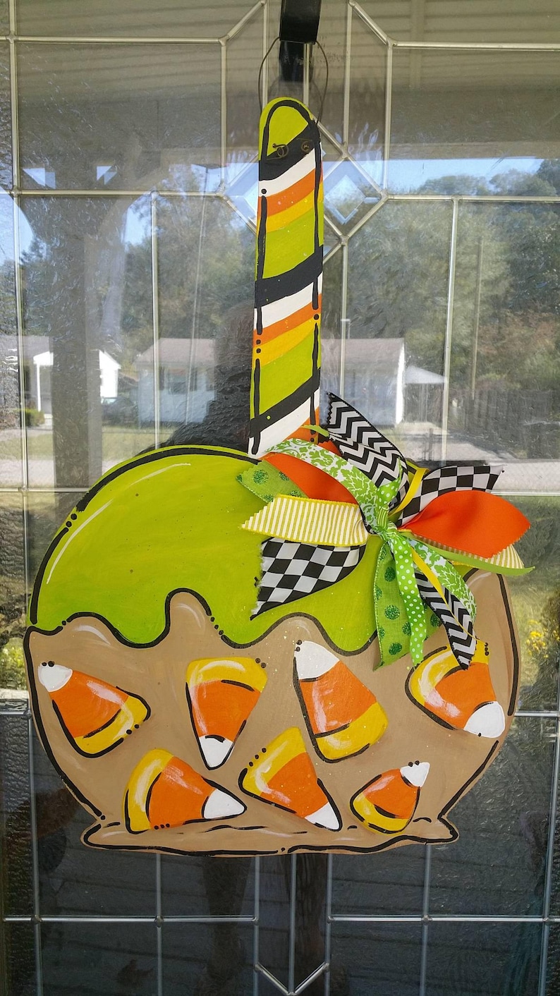 May include: A wooden Halloween-themed door hanger shaped like a candy corn container. The design features a green top, candy corn accents, and a colorful bow. The hanger is approximately 18 inches in height.