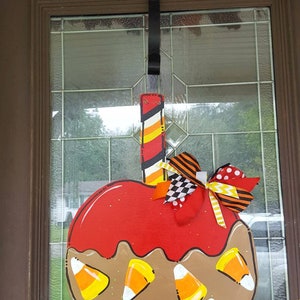 May include: A Halloween-themed wooden door hanger shaped like a caramel apple. The apple is red with brown caramel and candy corn accents. A decorative bow and a striped stick complete the design. The door hanger is suspended from a black ribbon.