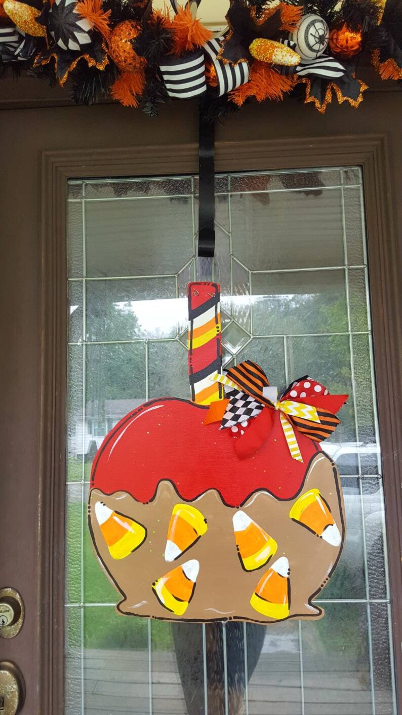 May include: A Halloween-themed wooden door hanger shaped like a caramel apple. The apple is red with brown caramel and candy corn accents. A decorative bow and a striped stick complete the design. The door hanger is suspended from a black ribbon.